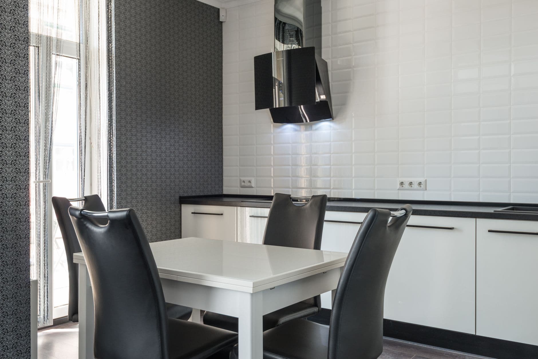 Interior of modern black and white kitchen furniture with white cupboards and diner table with black chairs near white tiled wall